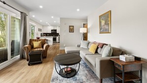 Ein modernes Wohnzimmer mit Sofa, Sessel und Couchtisch, im Hintergrund eine Küche und ein Balkon mit Blick.