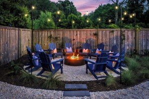 A backyard with a fire pit surrounded by Adirondack chairs, a wooden fence, and string lights.