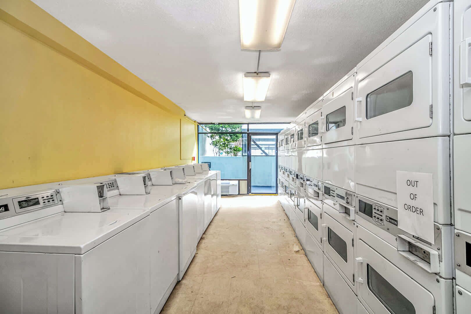 A laundry facility with rows of washing machines and dryers, a yellow wall, and a blue door leading outside.