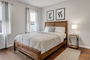 A bedroom with a wooden bed, a bedside table, a lamp, and two wall art pieces. - Cozy 3BR Medford l Minutes to Downtown lSleeps 6 (Medford)