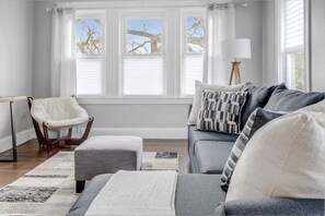 A living room with a grey sofa, a white chair, a lamp, and a window with blinds. - Cozy 3BR Medford l Minutes to Downtown lSleeps 6 (Medford)