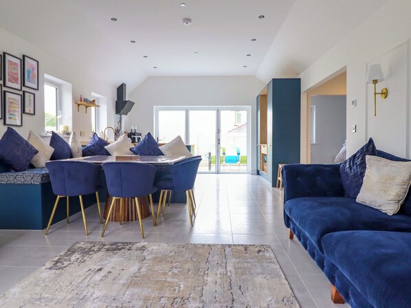 A modern living room with a blue sofa, a dining area with blue chairs and a table, and a large window providing natural light. - Kingfisher (Llanbedr)
