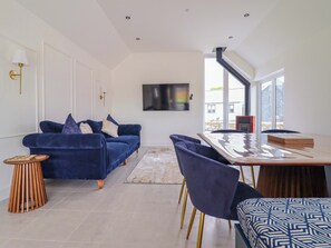 A modern living room with a blue sofa, a wooden side table, a large window, and a dining area with a wooden table and chairs. - Kingfisher (Llanbedr)