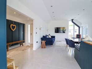 A spacious living area with a dark blue sofa, a wooden bench, a dining table with chairs, and a television mounted on the wall. - Kingfisher (Llanbedr)