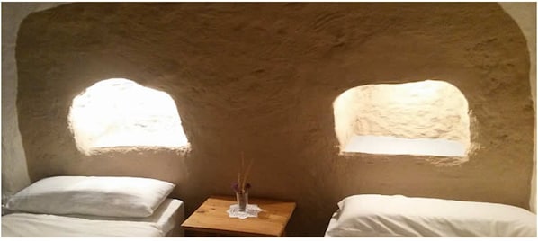 A room with two beds, a small table, and two windows with natural light.