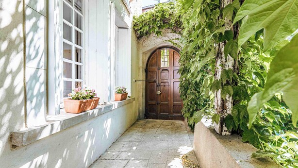 Un estrecho sendero de piedra que conduce a una puerta de madera, flanqueado por una pared blanca con ventanas y macetas.