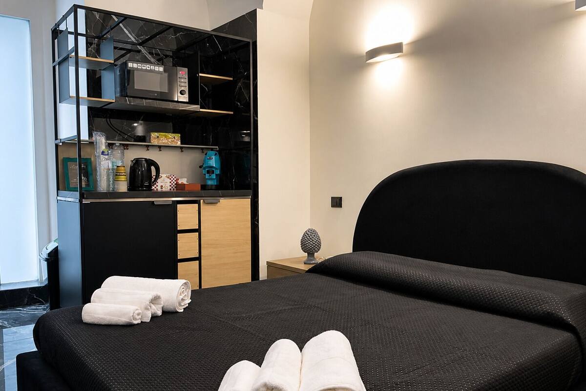 A hotel room with a bed, a kitchenette, and a small table with a lamp.