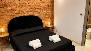 Moderni hotellihuone, jossa on tiiliseinä, musta sänky, kaksi yöpöytävalaisinta ja valkoinen pyyhe sängyllä.
