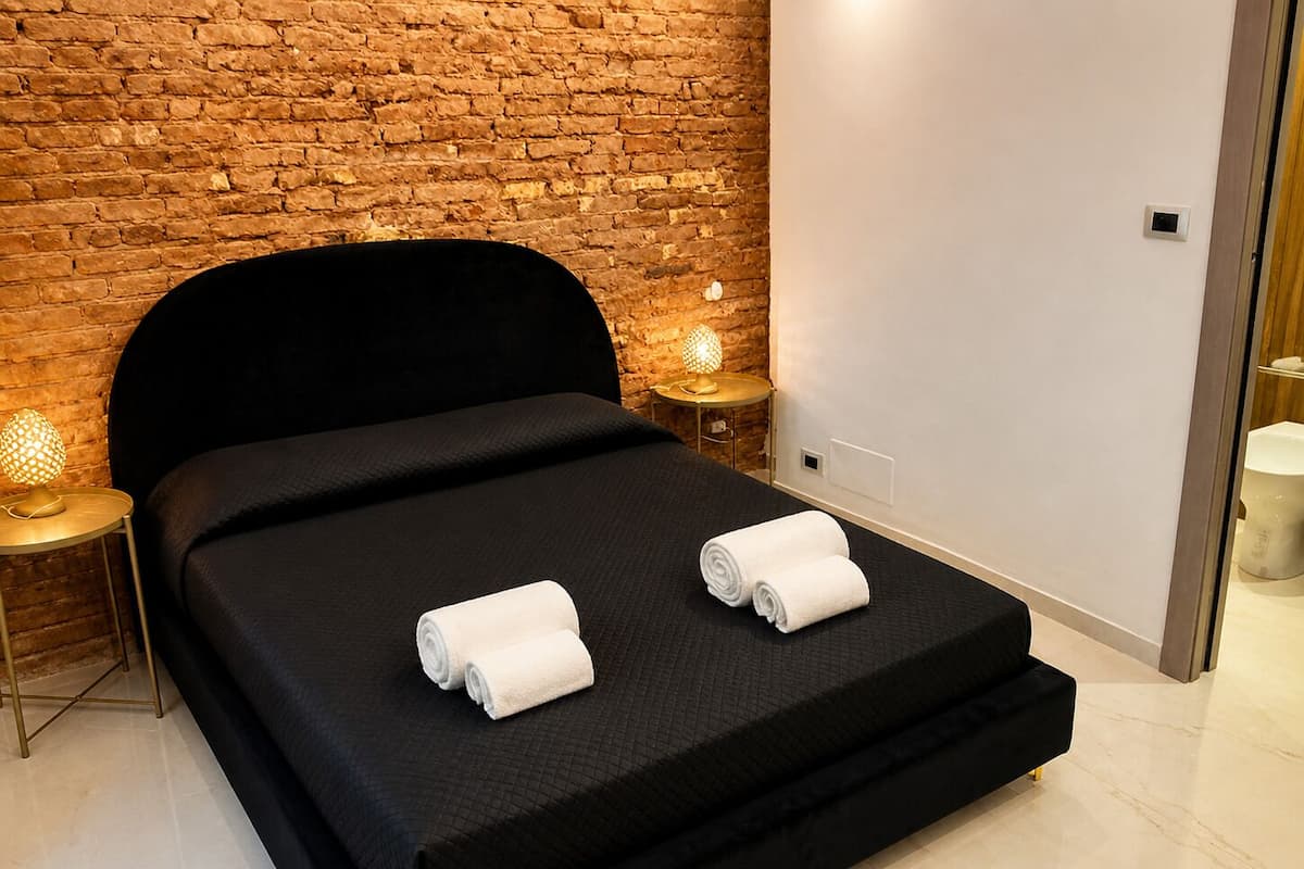 A modern hotel room with a brick wall, a black bed, two bedside lamps, and a white towel on the bed.