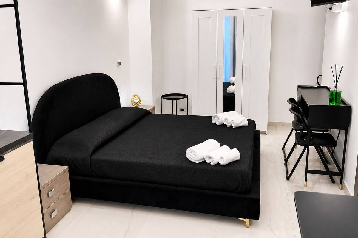 A modern hotel room with a black bed, a desk with a chair, a wardrobe, and an air conditioning unit.