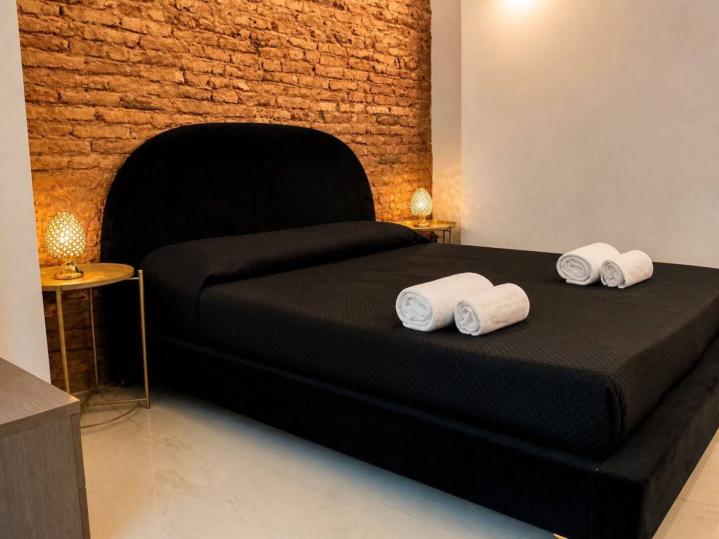 Un letto rifatto con cura, una testiera nera, due asciugamani bianchi, una parete in mattoni e due lampade da comodino.