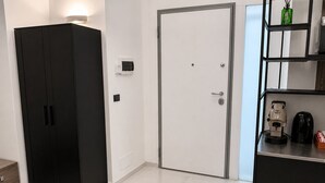 Hotellihuoneessa on musta vaatekaappi, valkoinen ovi, kahvinkeitin ja hylly, jonka päällä on vihreä pullo.