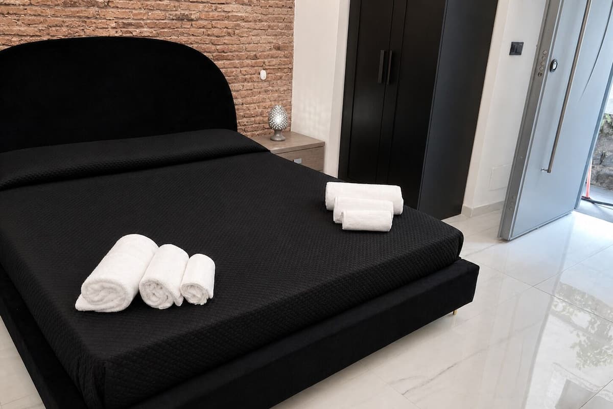 A modern hotel room with a black bed, a brick wall, a wooden nightstand, and a black wardrobe.