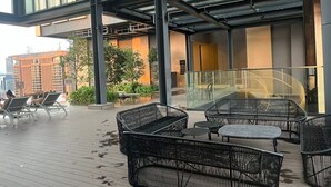 An outdoor seating area with wicker furniture, a glass railing, and a view of a building with a sign. - Luxury Suite Lucentia BBCC Infinity Pool (Kuala Lumpur)