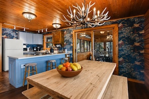 A wooden interior with a central island, a chandelier, and a dining table with a fruit bowl.