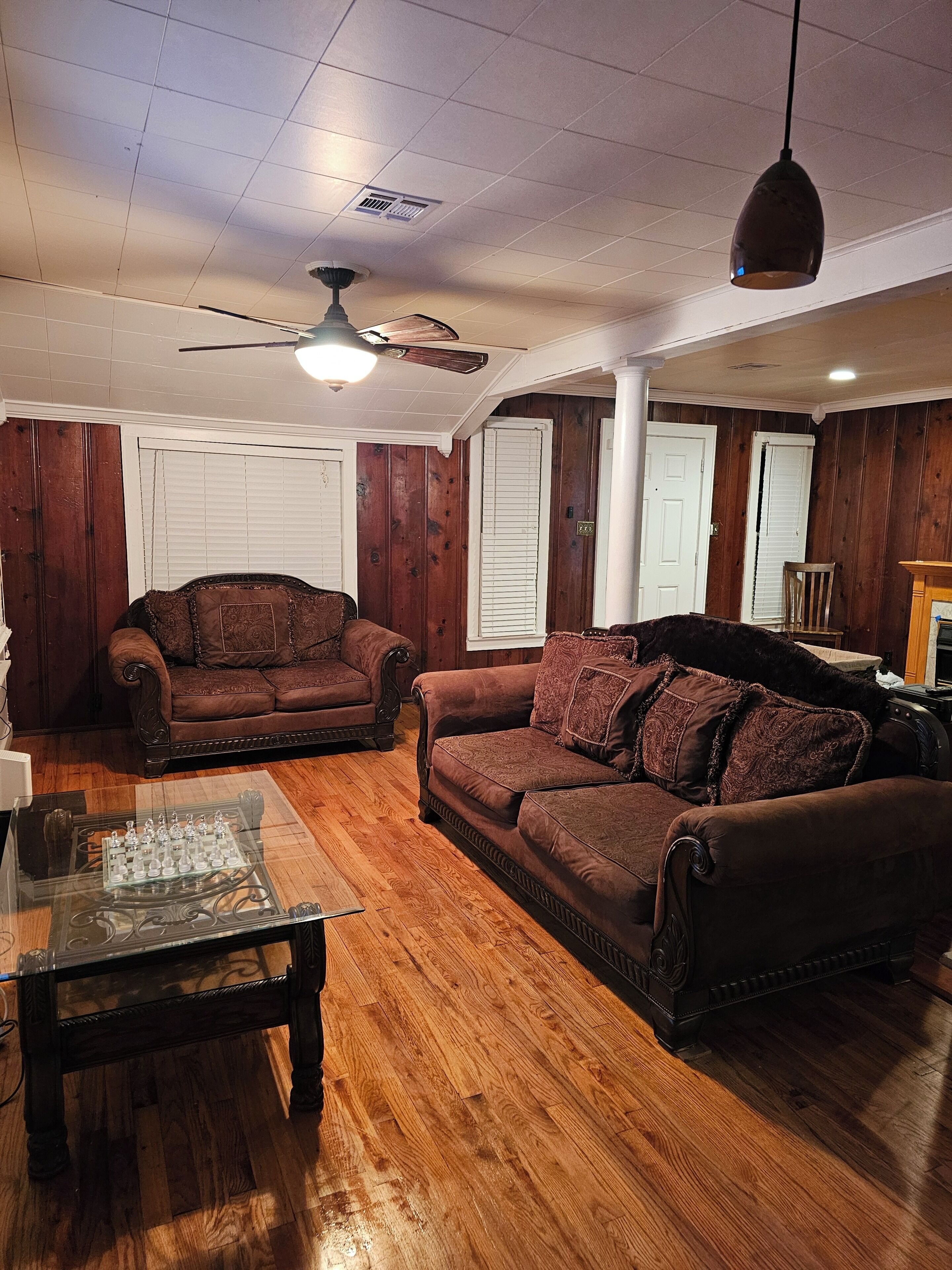 A living room with wooden paneling, two brown sofas, a glass coffee table, and a ceiling fan.