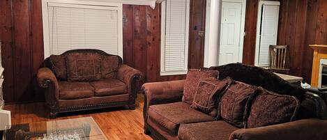 A living room with wooden paneling, two brown sofas, a glass coffee table, and a ceiling fan.