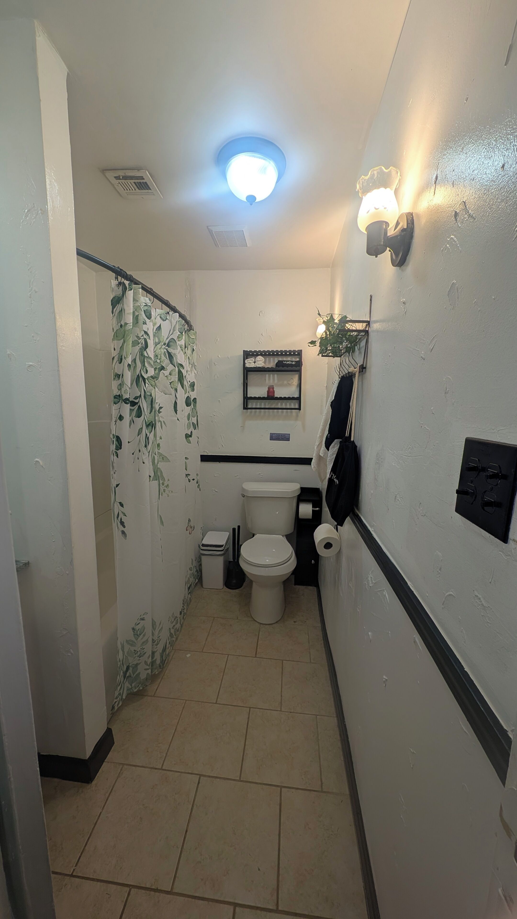 Un baño pequeño con inodoro, cortina de ducha, estante con artículos de tocador e iluminación.