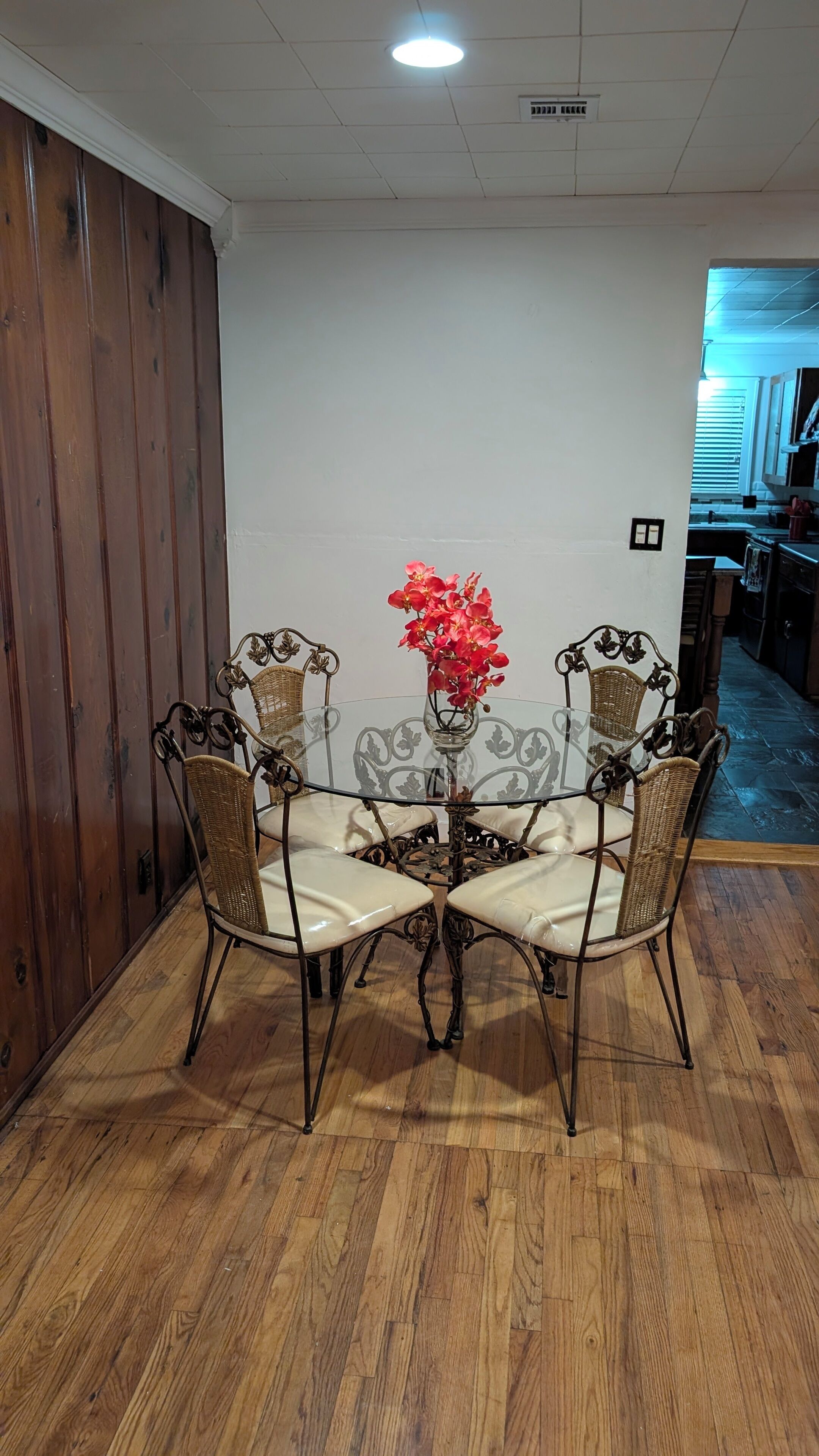 Un comedor con una mesa de vidrio rodeada de sillas de mimbre, un florero con flores y paneles de madera en una pared.