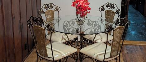Un comedor con una mesa de vidrio rodeada de sillas de mimbre, un florero con flores y paneles de madera en una pared.