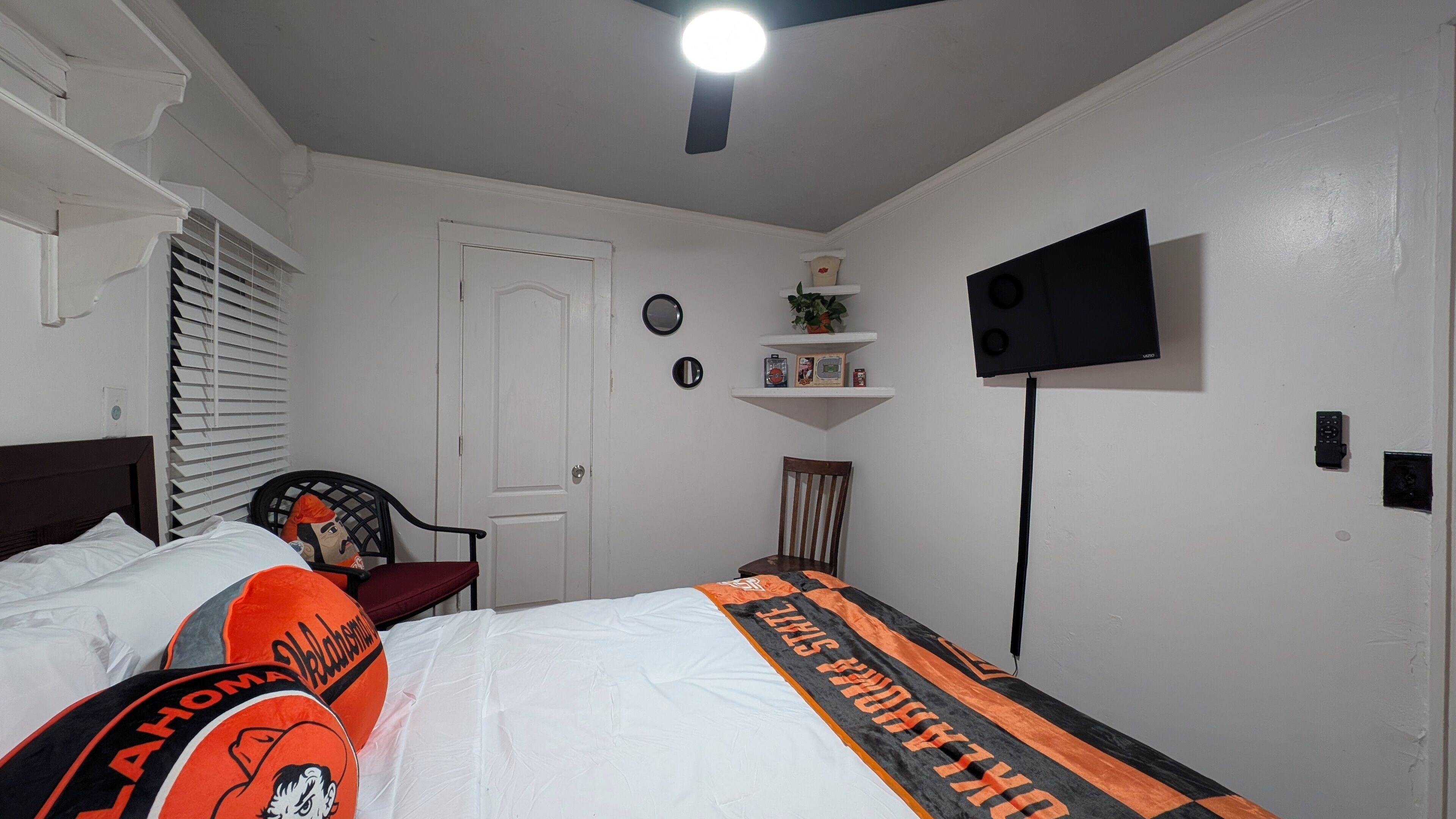 Un dormitorio con una cama, una silla, un televisor montado en la pared, una puerta y una ventana con persianas.
