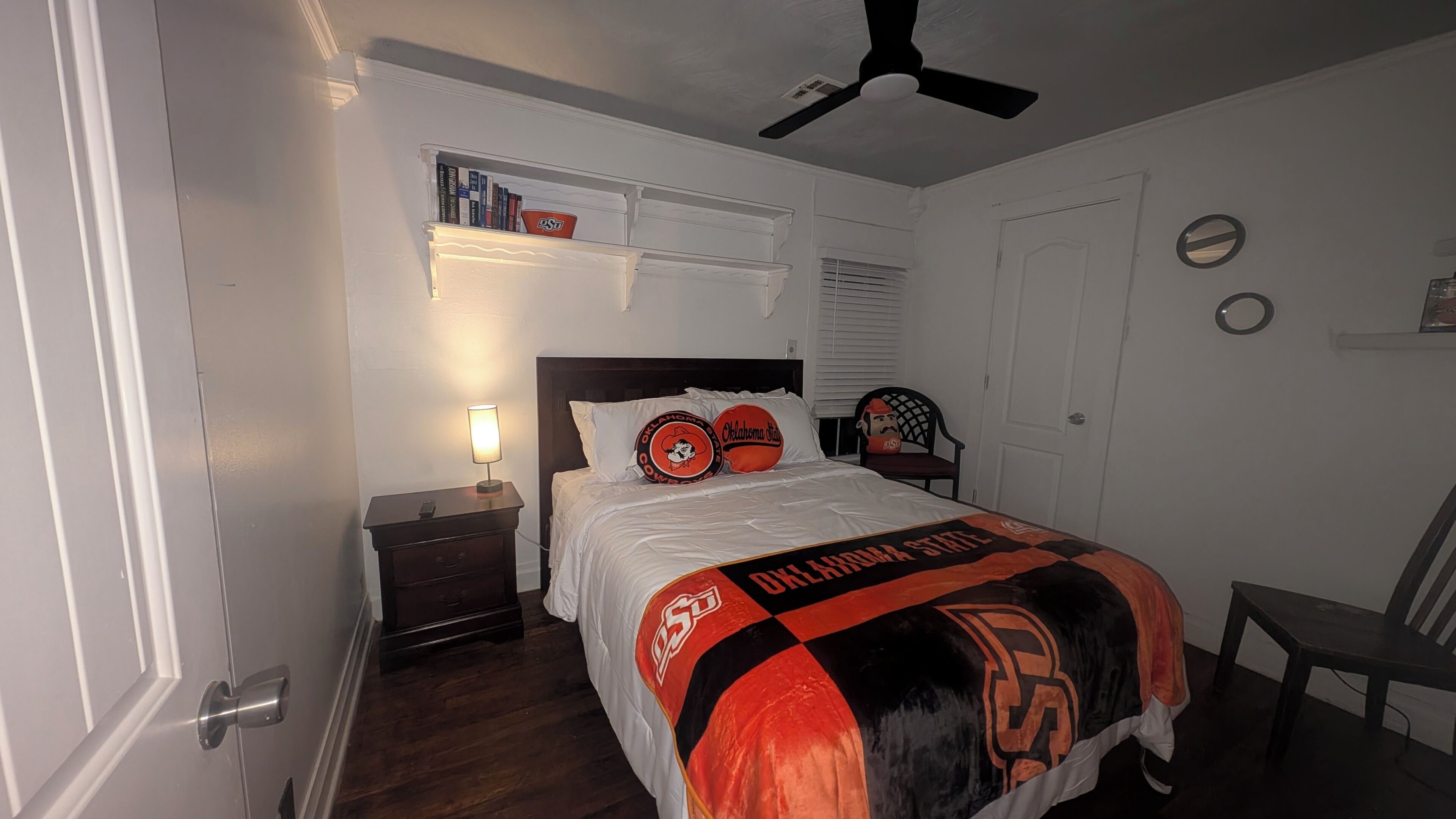 Een slaapkamer met een bed, nachtkastje, plafondventilator, plank met boeken en een deur.