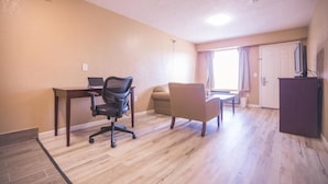 A room with a desk, office chair, sofa, and a television on a stand. - Crossroads Hotel and Suites Irving (Irving)