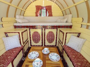 A cozy interior with a wooden table set for tea, cushioned seating, and a window with curtains. - The Bowtop Gypsy Caravan (Eye)