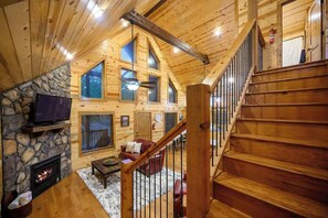 Ein Holzhütten-Innenraum mit Kamin aus Stein, einem Fernseher, einer Treppe und einer gemütlichen Sitzecke.