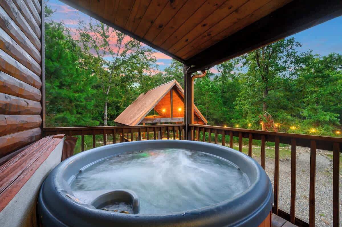 A hot tub on a wooden deck with a view of a cabin in a forest.