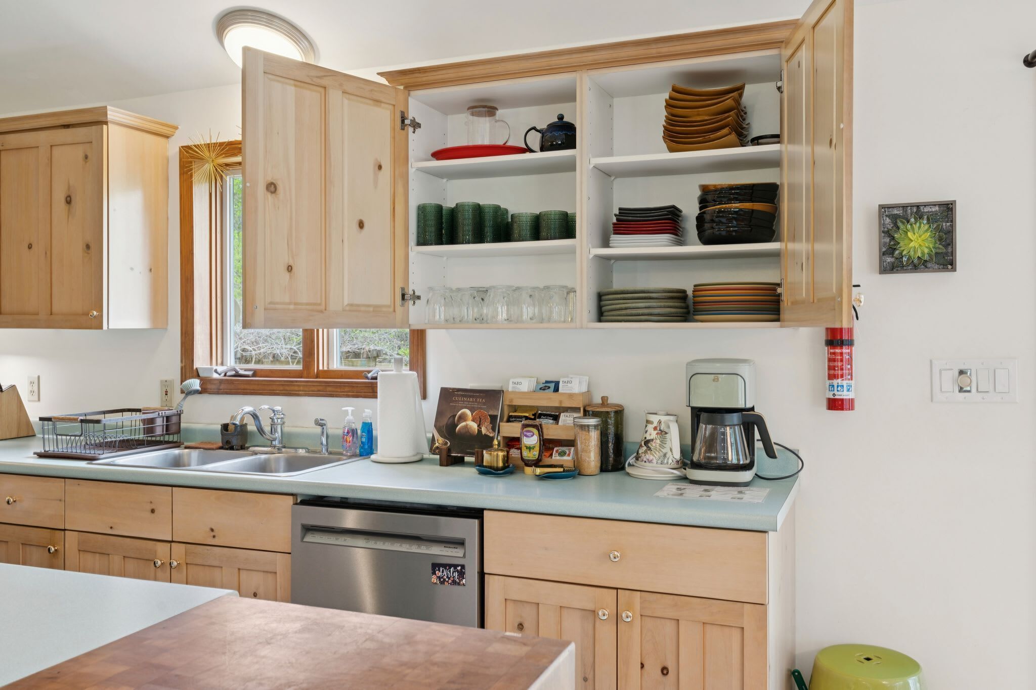 Une cuisine avec des meubles de rangement en bois, un lave-vaisselle, une machine à café et divers ustensiles de cuisine disposés sur le plan de travail.