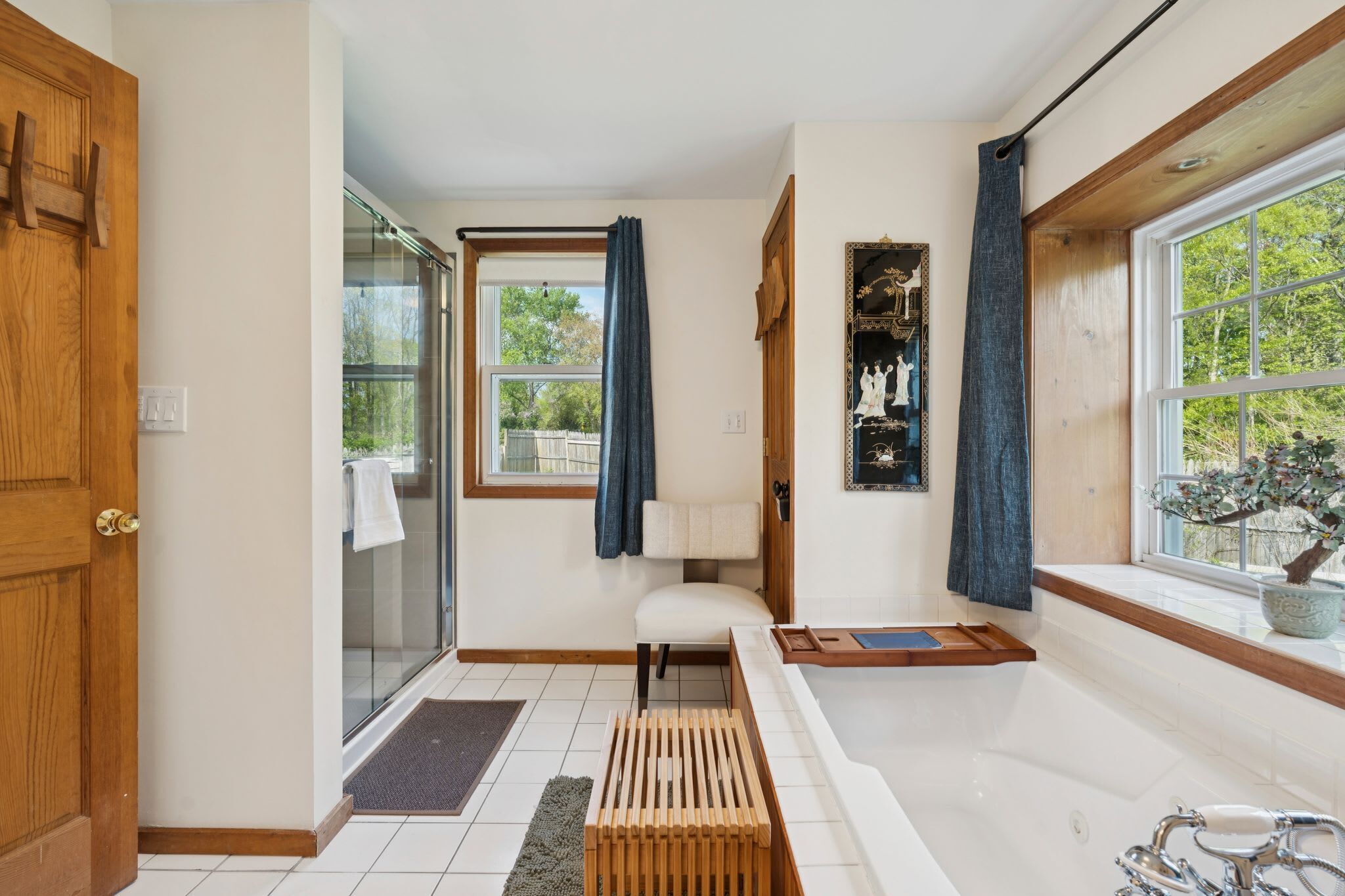 Ein Badezimmer mit einer Badewanne, einer Dusche, einer hölzernen Sitzbank und einem Fenster mit Blick auf Bäume.