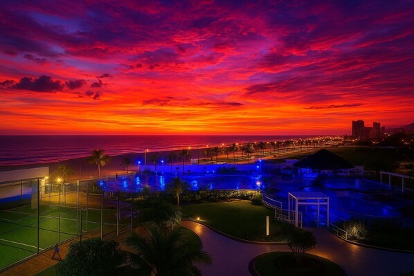 A resort with a pool area, palm trees, and a view of the ocean at sunset.
