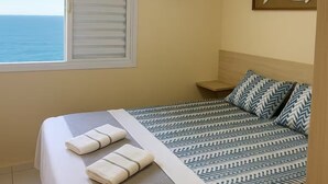 Un dormitorio con cama, ventilador de techo, ventana con vista al mar y dos adornos de pared.