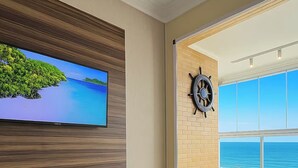 Una sala de estar moderna con un televisor de pantalla plana montado en una pared de paneles de madera, un control remoto sobre un sofá gris, un adorno con forma de timón en la pared y un amplio ventanal con vistas al mar.