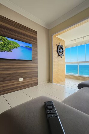 A modern living room with a flat-screen TV mounted on a wooden panel wall, a large window with a view of the ocean, and a remote control on a grey sofa.