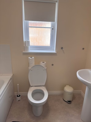 A bathroom with a toilet, a window, a sink, and a trash can.