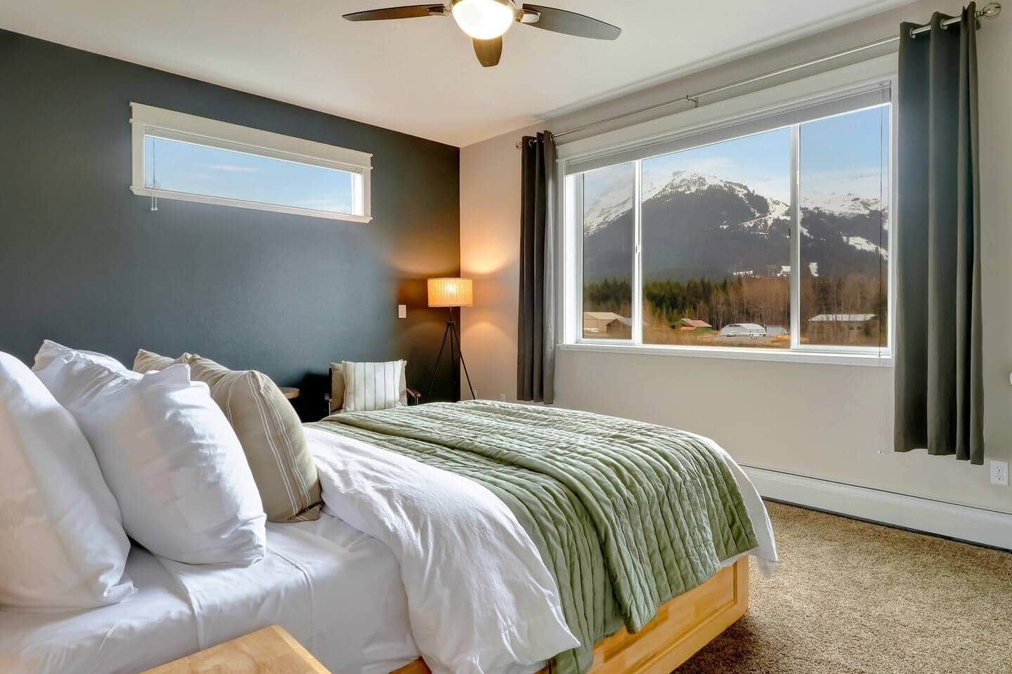 A bedroom with a bed, a standing lamp, a ceiling fan, and a large window with a mountain view.