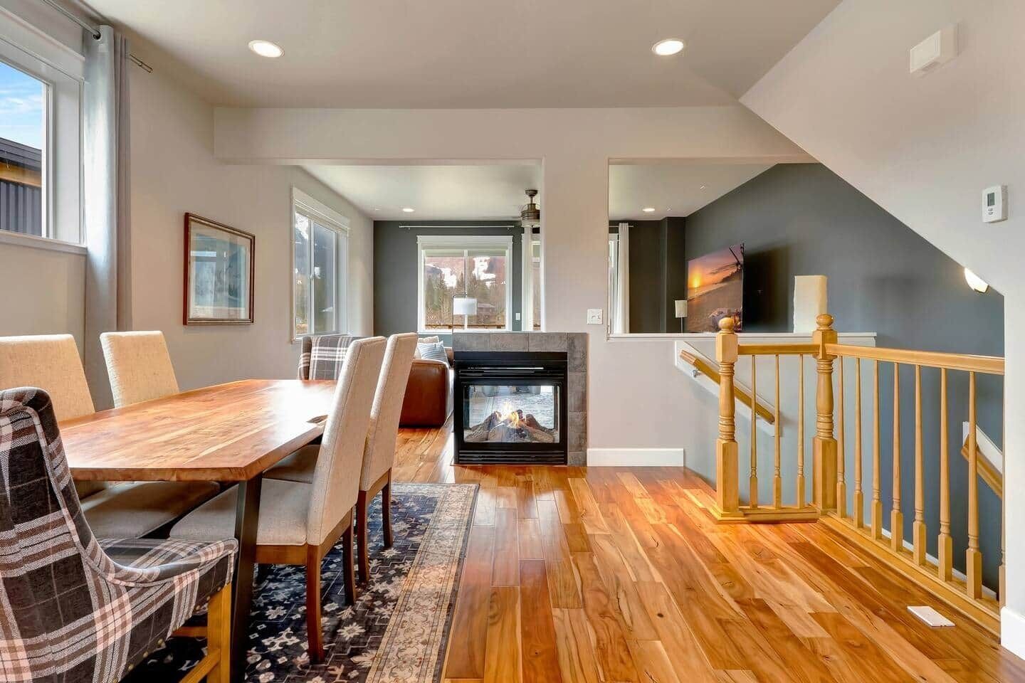 A modern living room with a fireplace, wooden flooring, a dining area with chairs, and a staircase with a railing.