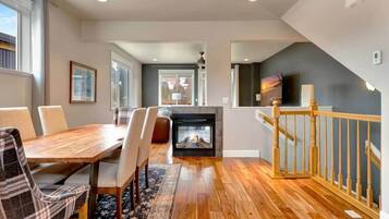 A modern living room with a fireplace, wooden flooring, a dining area with chairs, and a staircase with a railing.