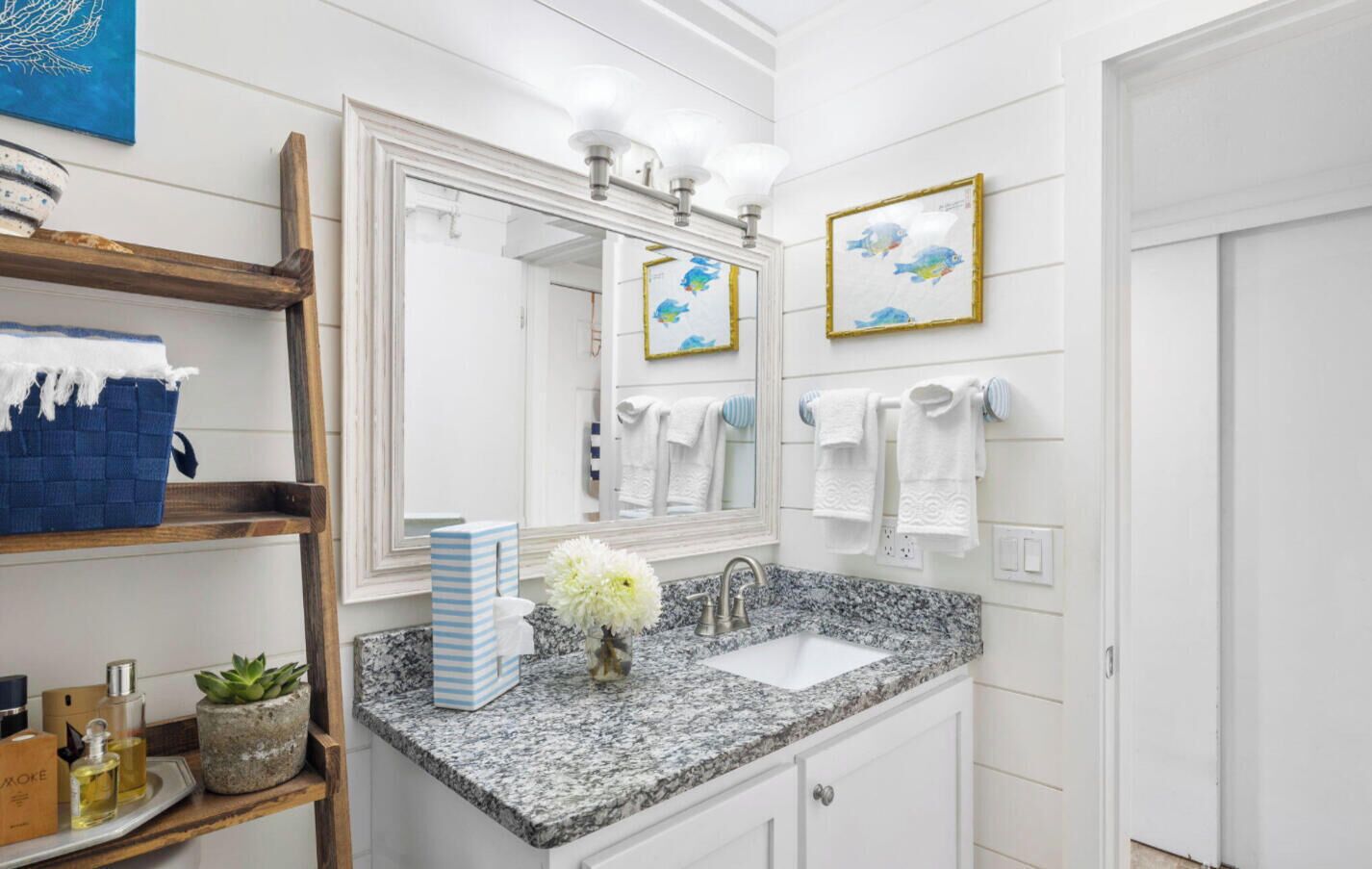 A bathroom with a granite countertop, a sink, a mirror with lights, a towel rack, and a shelf with towels and a blue basket.