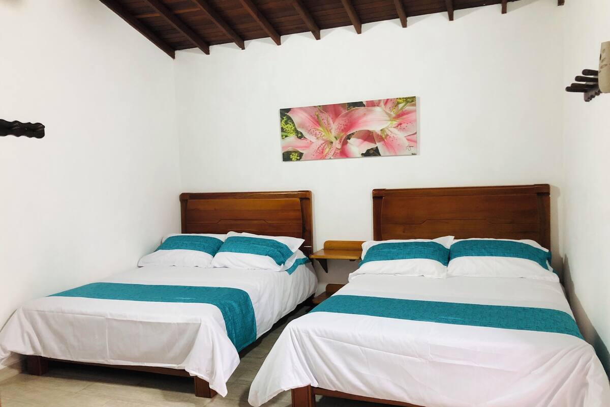 Una habitación con dos camas de madera, ropa de cama blanca, detalles color turquesa, un cabecero de madera, un banco de madera y un cuadro floral en la pared.