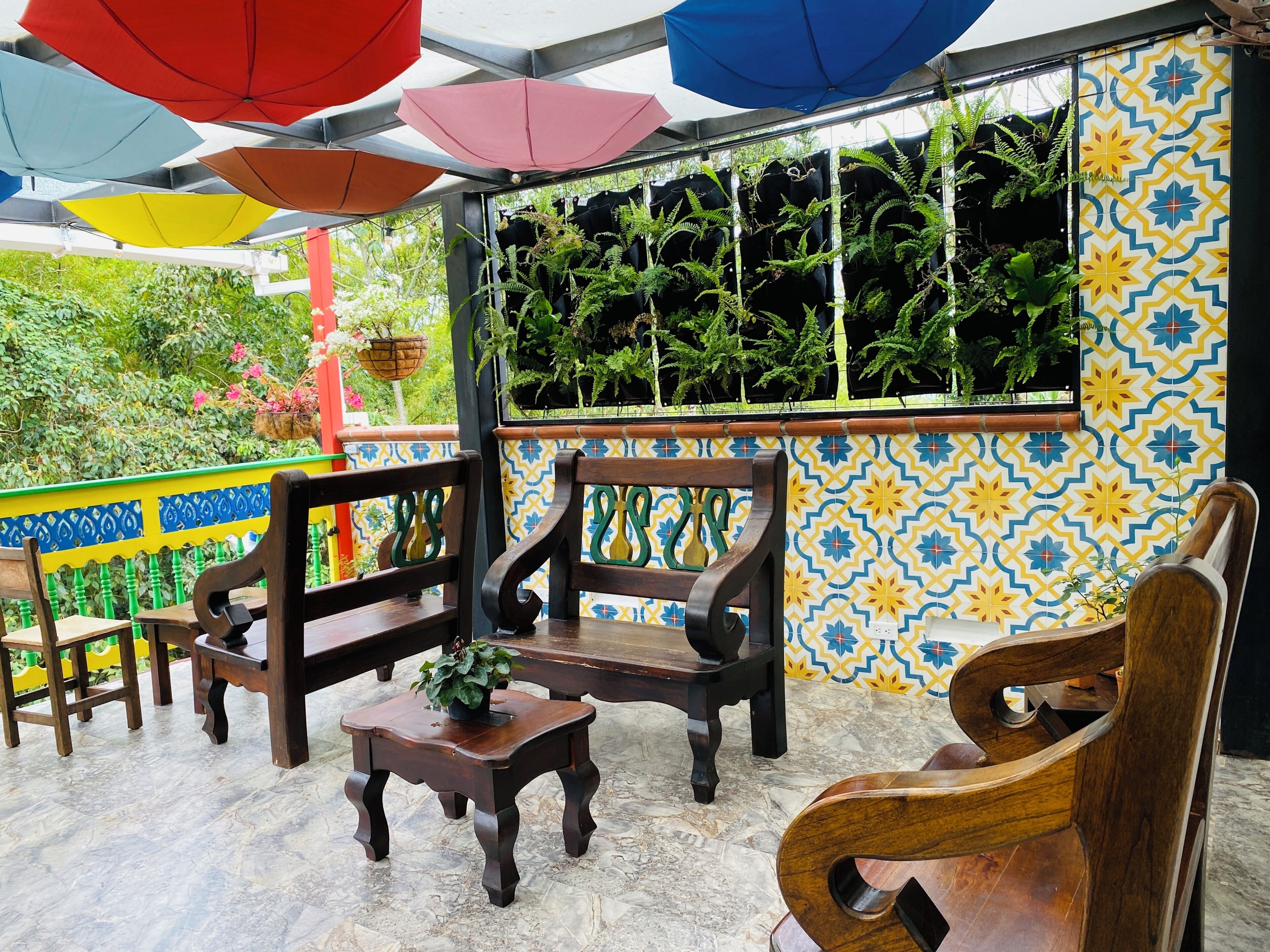 Eine Terrasse mit Holzmöbeln, einer Wand mit geometrischem Muster, hängenden Pflanzen und bunten Sonnenschirmen.