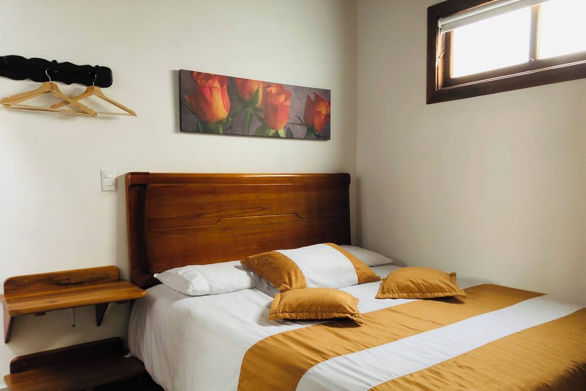 Un dormitorio con cabecera de madera, cama con ropa de cama blanca y mostaza, mesita de noche, banco de madera y un cuadro de flores en la pared.