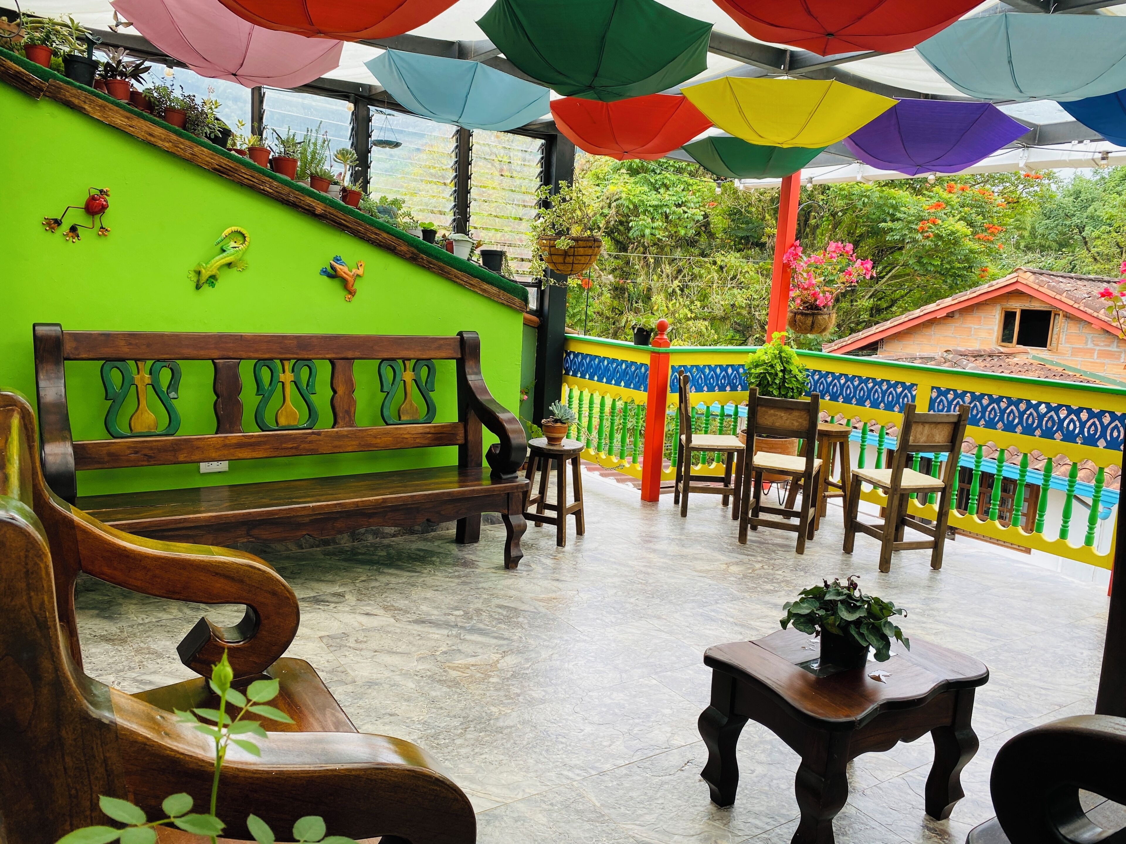 A patio with a wooden bench, chairs, and tables, surrounded by colorful walls and potted plants.