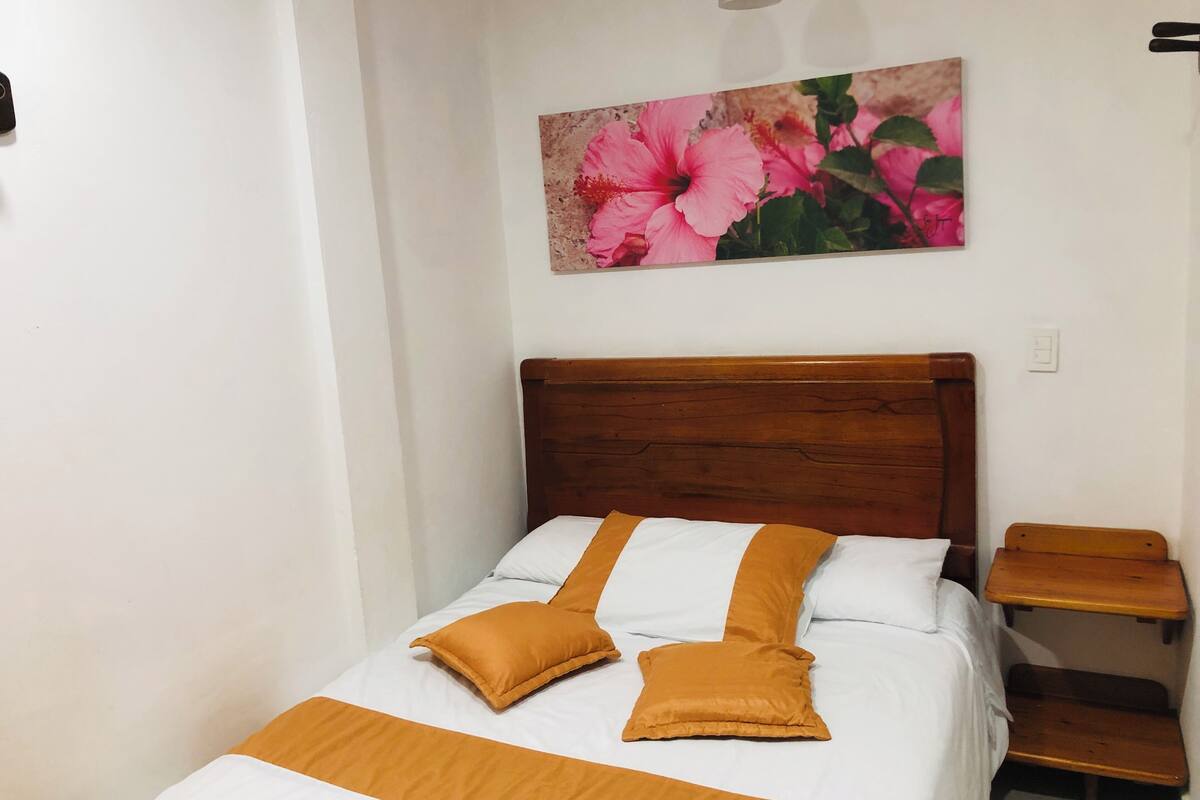 Un dormitorio con cabecera de madera, ropa de cama blanca y un cuadro de flores rosadas en la pared.