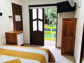 Une pièce avec un lit, un bureau en bois, une porte donnant sur l’extérieur et un ventilateur fixé au mur.