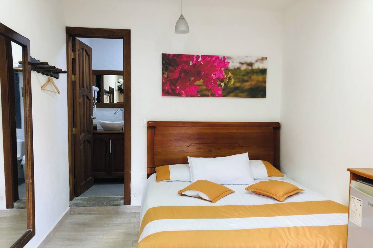 Un dormitorio con cama de madera, pared blanca, un cuadro de flores y un baño visible a través de una puerta abierta.