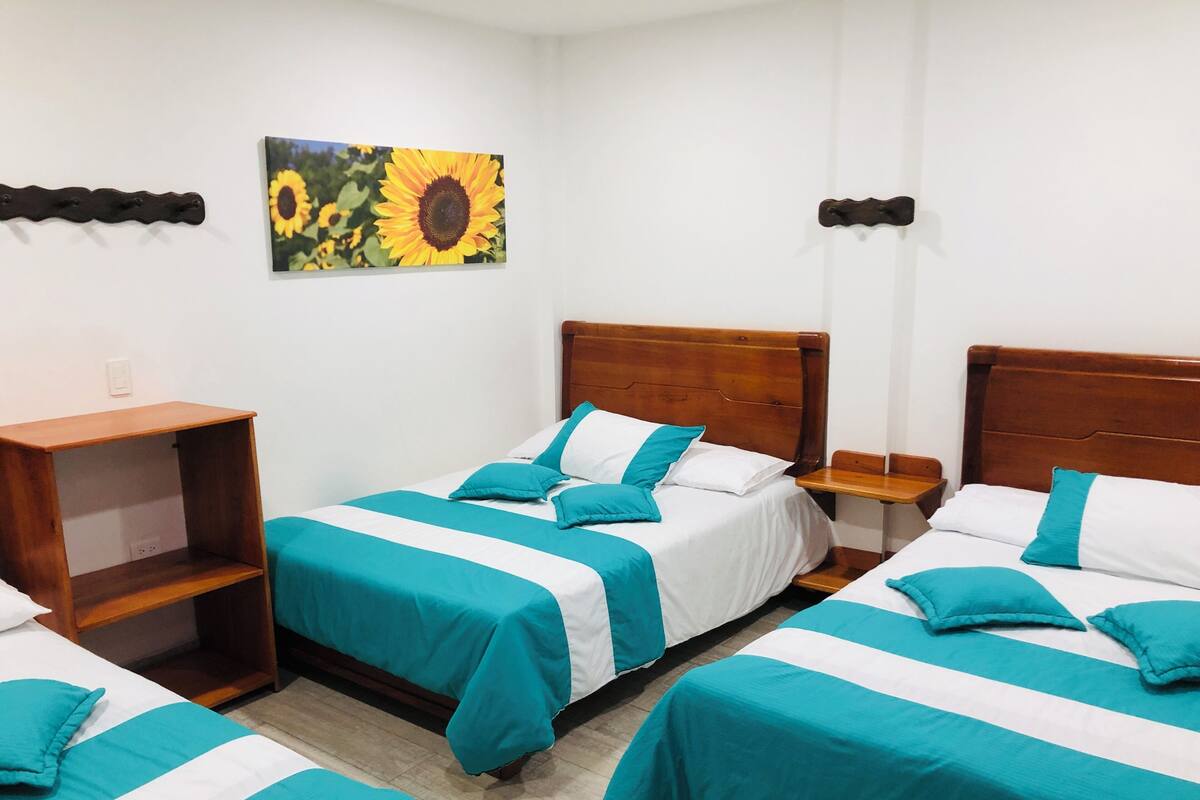 Habitación de hotel con dos camas individuales, un cabecero de madera, una mesita de noche pequeña de madera, una estantería y un cuadro de girasoles.