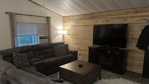 A living room with a sofa, a coffee table, a TV on a stand, a lamp, and a window with blinds. - Mountain House - Mt. Rainier Cabin - Hot Tub, Fire Pit, Pet Friendly. (Ashford)